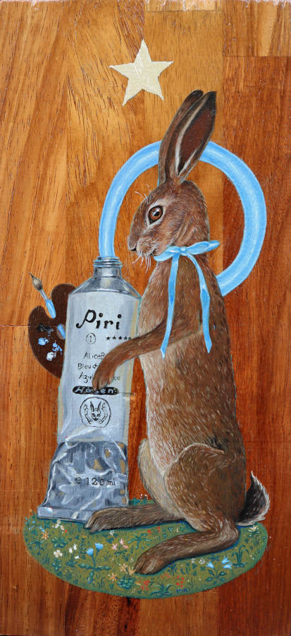 Block of red wood with a standing hare wearing a blue bow painted on it. The hare is holding onto a giant paint tube that is squirting blue paint in a circle. The hare is also holding a palette and brush and is standing on a circle mound of grass and flowers in a Mille Fleur style. Above the hare is a silver star.